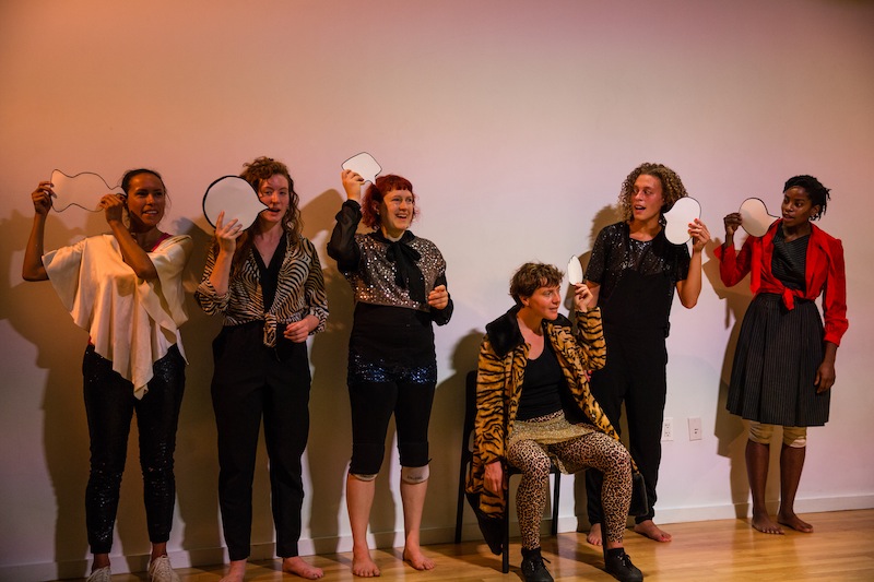 The cast, in wild animal prints and outlandish ensembles, hold up card board word bubbles
